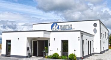 Modernes, zweigeschossiges Firmengebäude mit weißer Fassade und flachem Dach. Über dem Eingang prangt das Logo ‚Martino Heizung-Sanitär-Klima‘. Vor dem Gebäude befinden sich gepflegte Beete mit Pflanzen sowie ein gepflasterter Parkplatz. Der Himmel ist teils bewölkt, aber hell.