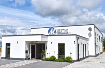 Modernes, zweigeschossiges Firmengebäude mit weißer Fassade und flachem Dach. Über dem Eingang prangt das Logo ‚Martino Heizung-Sanitär-Klima‘. Vor dem Gebäude befinden sich gepflegte Beete mit Pflanzen sowie ein gepflasterter Parkplatz. Der Himmel ist teils bewölkt, aber hell.