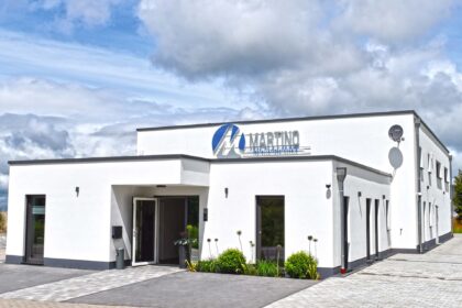 Martino Heizung Merzig Modernes, zweigeschossiges Firmengebäude mit weißer Fassade und flachem Dach. Über dem Eingang prangt das Logo ‚Martino Heizung-Sanitär-Klima‘. Vor dem Gebäude befinden sich gepflegte Beete mit Pflanzen sowie ein gepflasterter Parkplatz. Der Himmel ist teils bewölkt, aber hell.