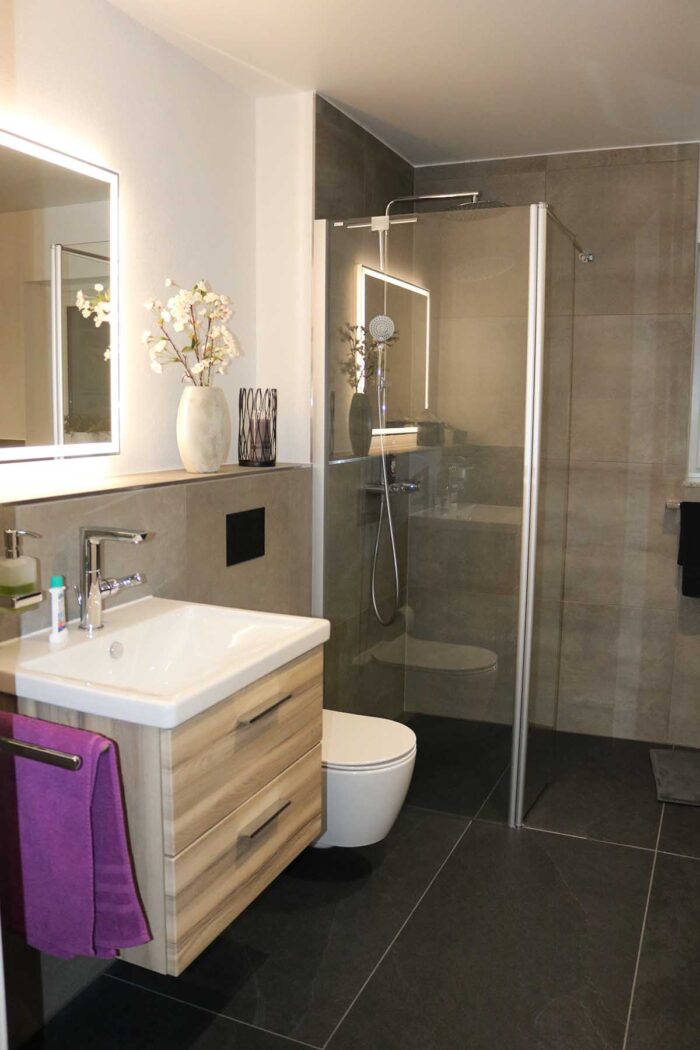 Modernes Badezimmer mit freistehender Badewanne, schwarzem Wandregal, großformatigen grauen Fliesen und sanfter, indirekter Beleuchtung.