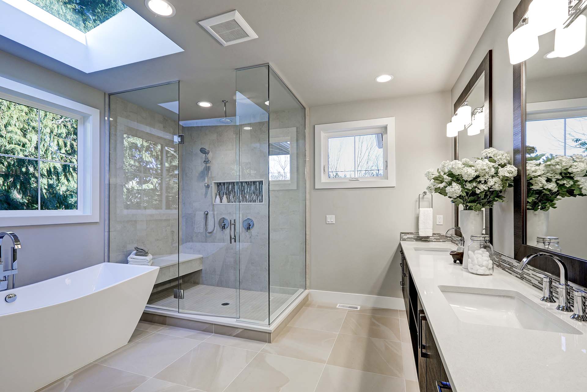 Modernes, helles Badezimmer mit großer, bodengleicher Glasdusche, freistehender Badewanne und Doppelwaschtisch mit zwei Spiegeln. Das Tageslicht fällt durch zwei Fenster und ein Oberlicht herein. Auf der Ablage stehen weiße Blumenvasen, Handtücher und Pflegeartikel. Der Raum wirkt sauber, stilvoll und einladend. Alles wurde von der Firma Martino aus Merzig saniert.